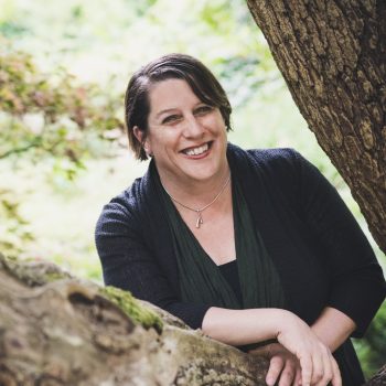 Image of Marina Psaros leaning on tree. She is smiling and looking at camera, wearing a black cardigan and black top.